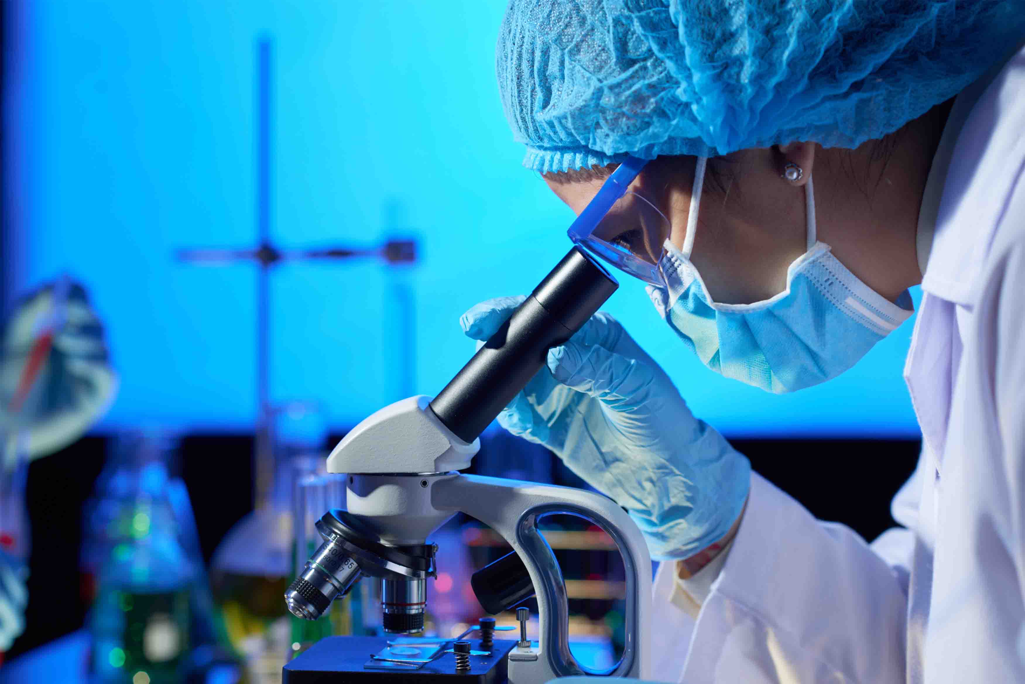 Personal de laboratorio analizando muestras en un microscopio; viste bata blanca y guantes de látex azules; las instalaciones y la iluminación muestran tonos azules.